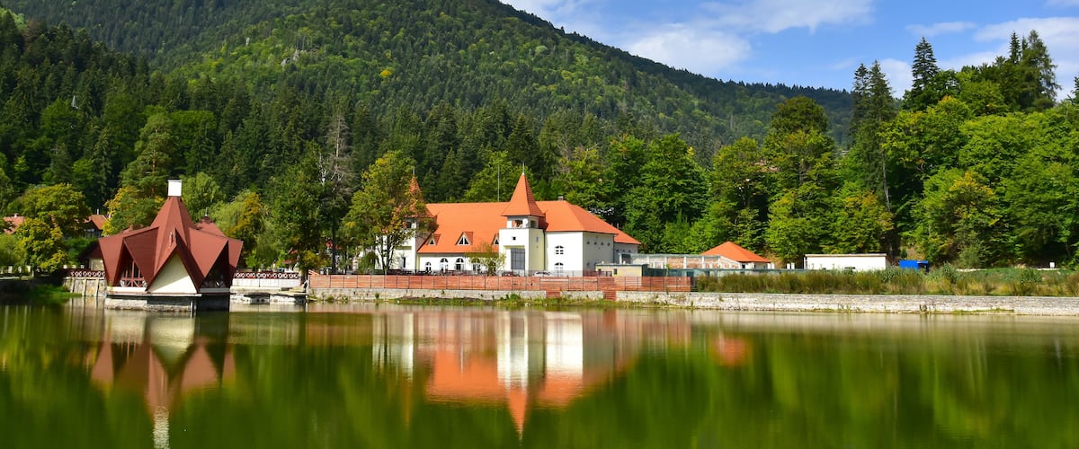 A lakeside view with a red-roofed building, a white building, and a forested hill.