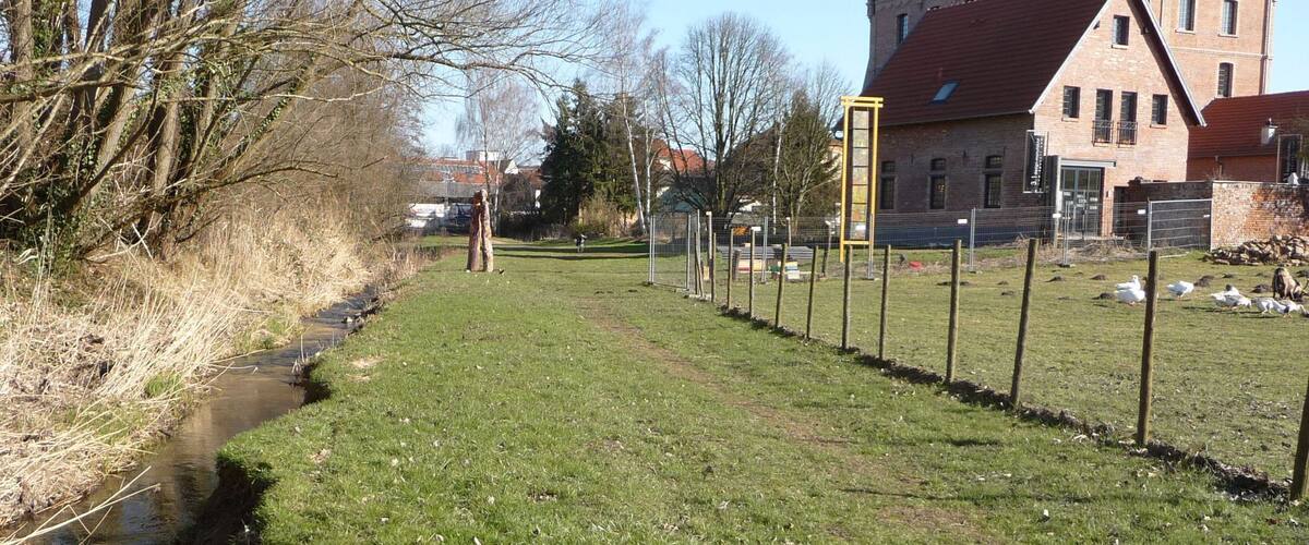 A grassy area with a small stream, a fence, and a building with a red roof in the background.