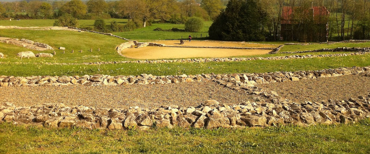 A historical site with stone walls and a circular enclosure, surrounded by green fields and trees.