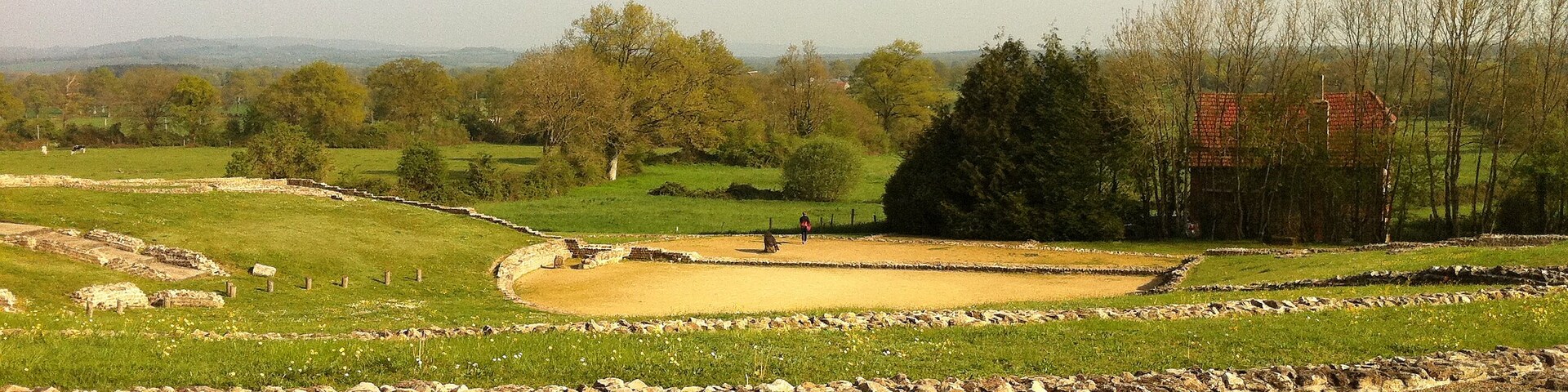 A historical site with stone walls and a circular enclosure, surrounded by green fields and trees.