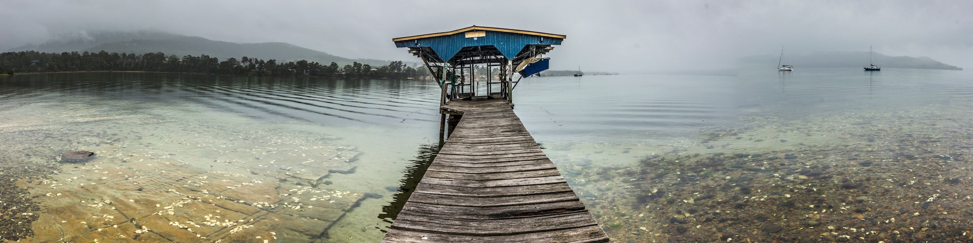 A wooden pier leading to a blue shelter by a calm lake with clear water and floating leaves.