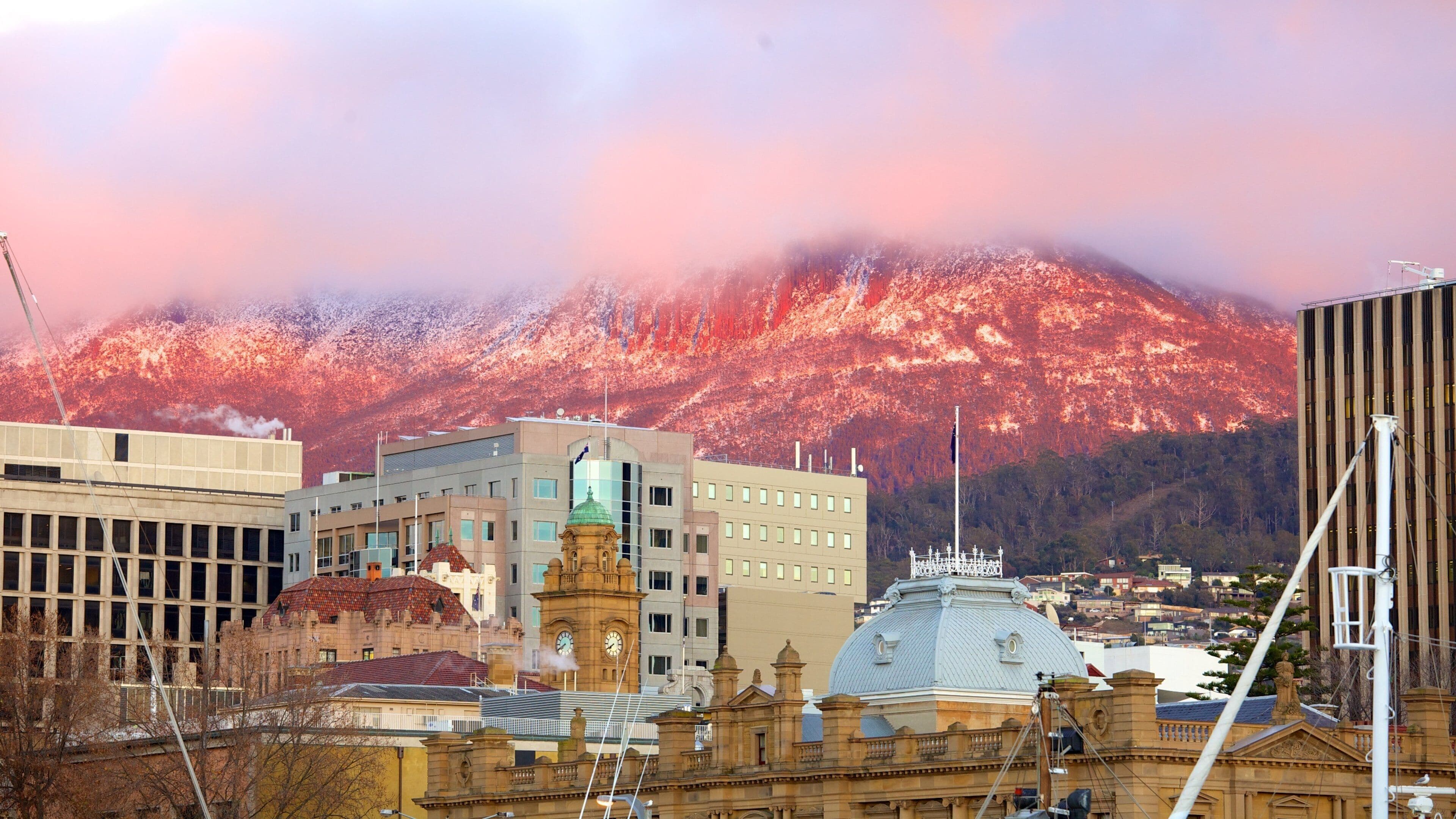 Un paisaje urbano con edificios modernos y una torre de reloj histórica, frente a un paisaje montañoso cubierto de nieve y un cielo rosado.