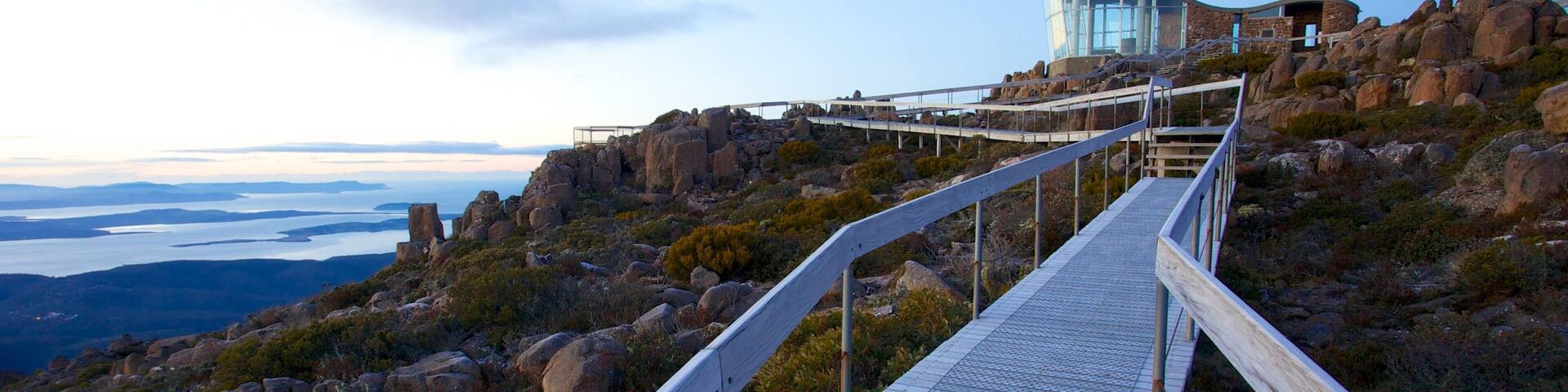 A wooden boardwalk leading to a glass building on a rocky hillside with a view of a body of water in the distance.