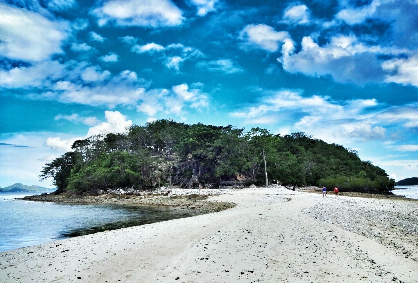 A beach with white sand, a small island with dense green trees, and a clear blue sky with scattered clouds.