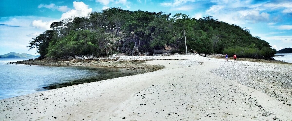 A beach with white sand, a small island with dense green trees, and a clear blue sky with scattered clouds.