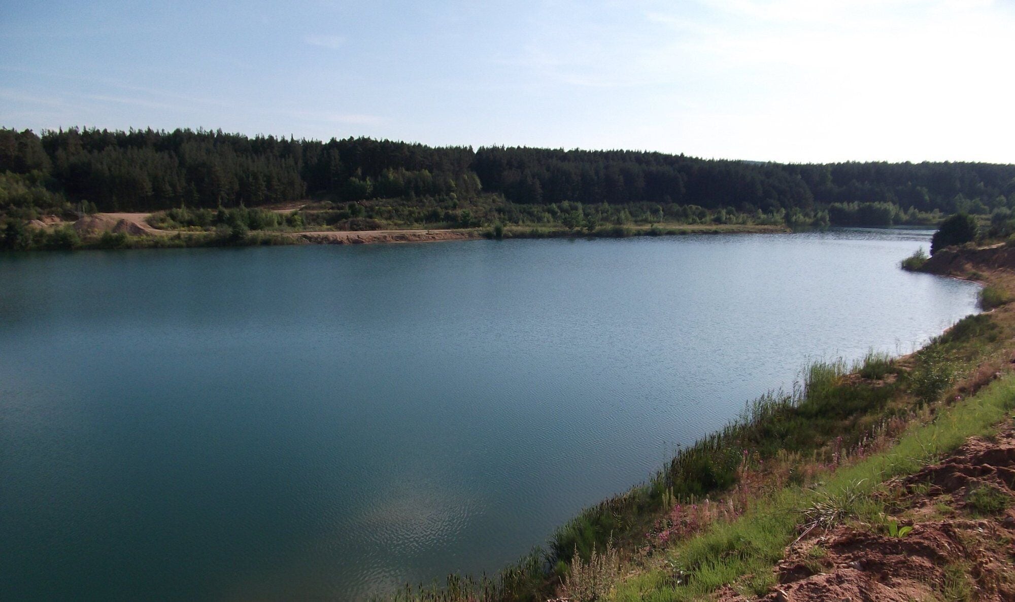 A calm lake surrounded by dense forest and a dirt path along the shore.