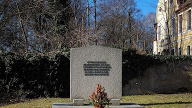 Une pierre tombale surmontée d’une croix, entourée d’arbres, avec un bâtiment visible au loin.