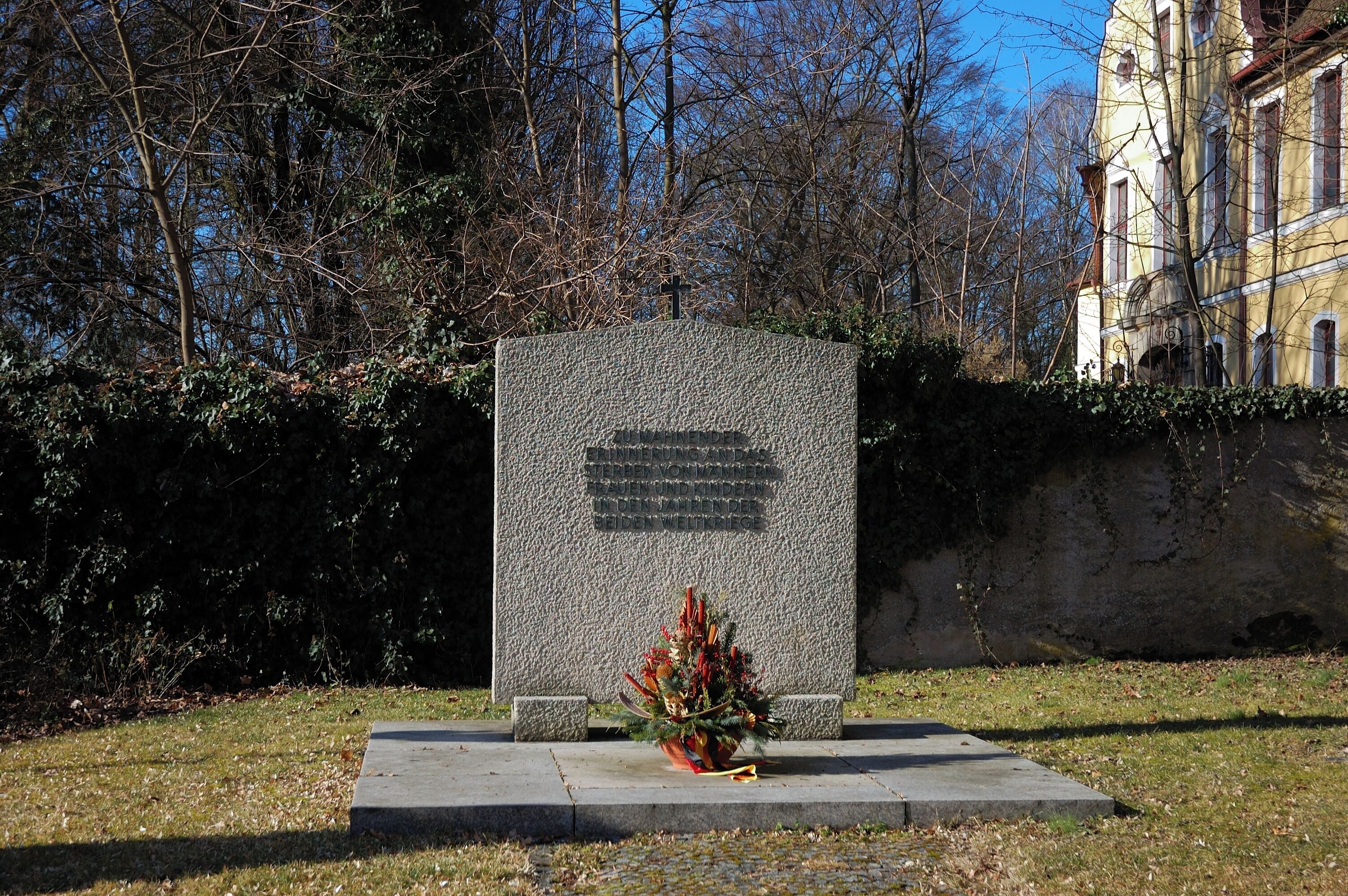 A gravestone with a cross, surrounded by trees and a building in the background.