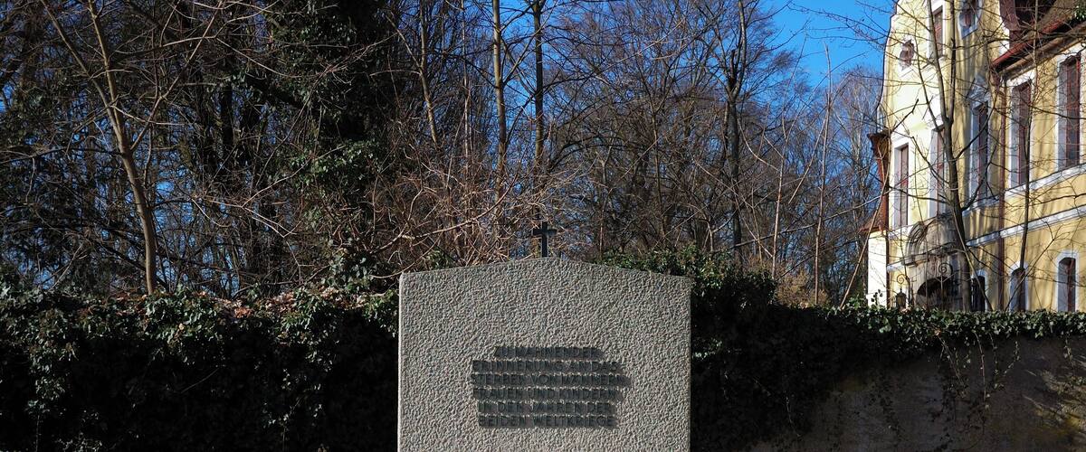 A gravestone with a cross, surrounded by trees and a building in the background.