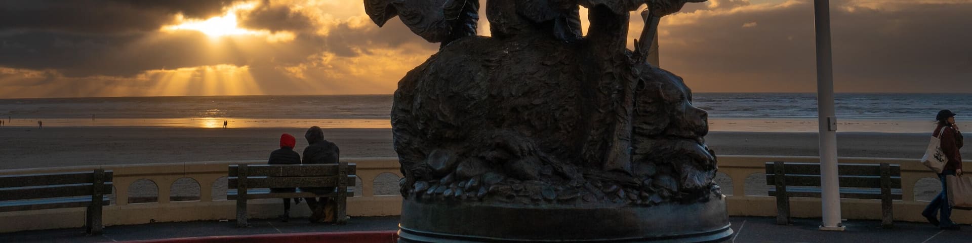 A statue of two figures with a flag, a bench, and a beach in the background.