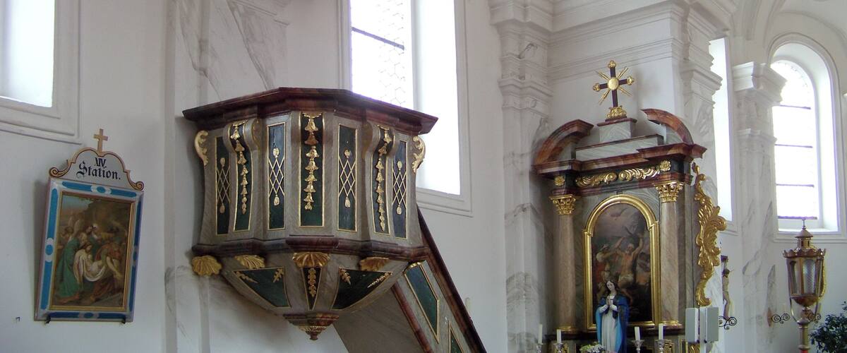 A church interior with ornate decorations, including a large wooden structure with gold accents and a painting of a religious scene.