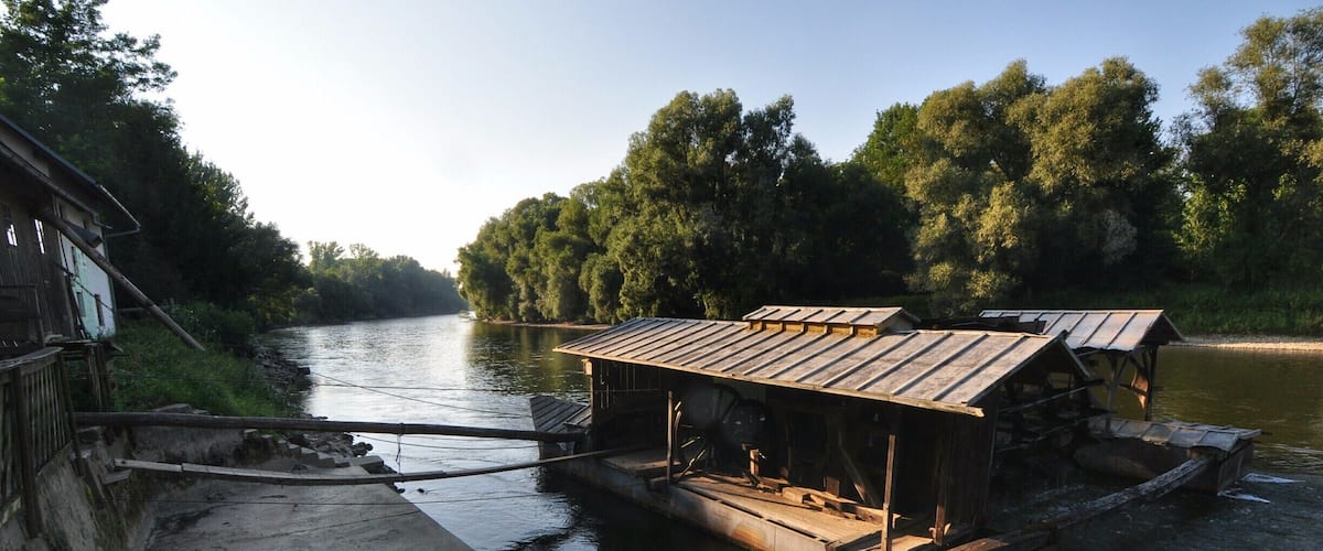 A wooden boat docked at a concrete pier with a metal roof, surrounded by trees and a calm river.