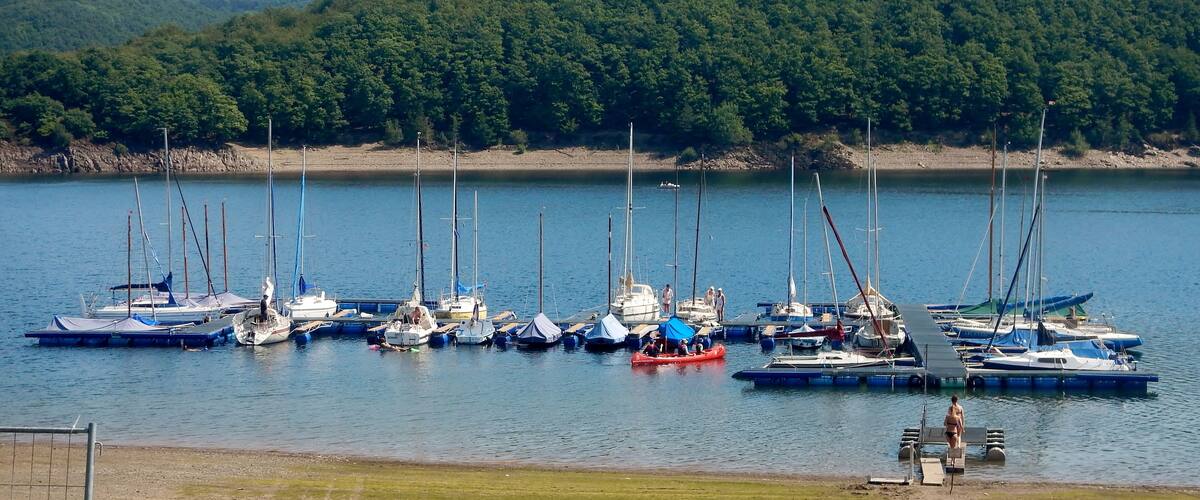 A marina with several sailboats docked along the shore, surrounded by a forested hillside.