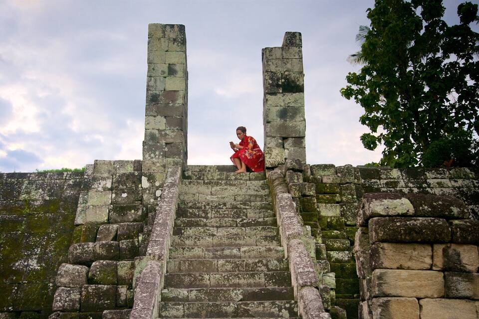 Una mujer con vestido rojo sentada sobre antiguos escalones de piedra, con paredes cubiertas de musgo y una palmera al fondo.