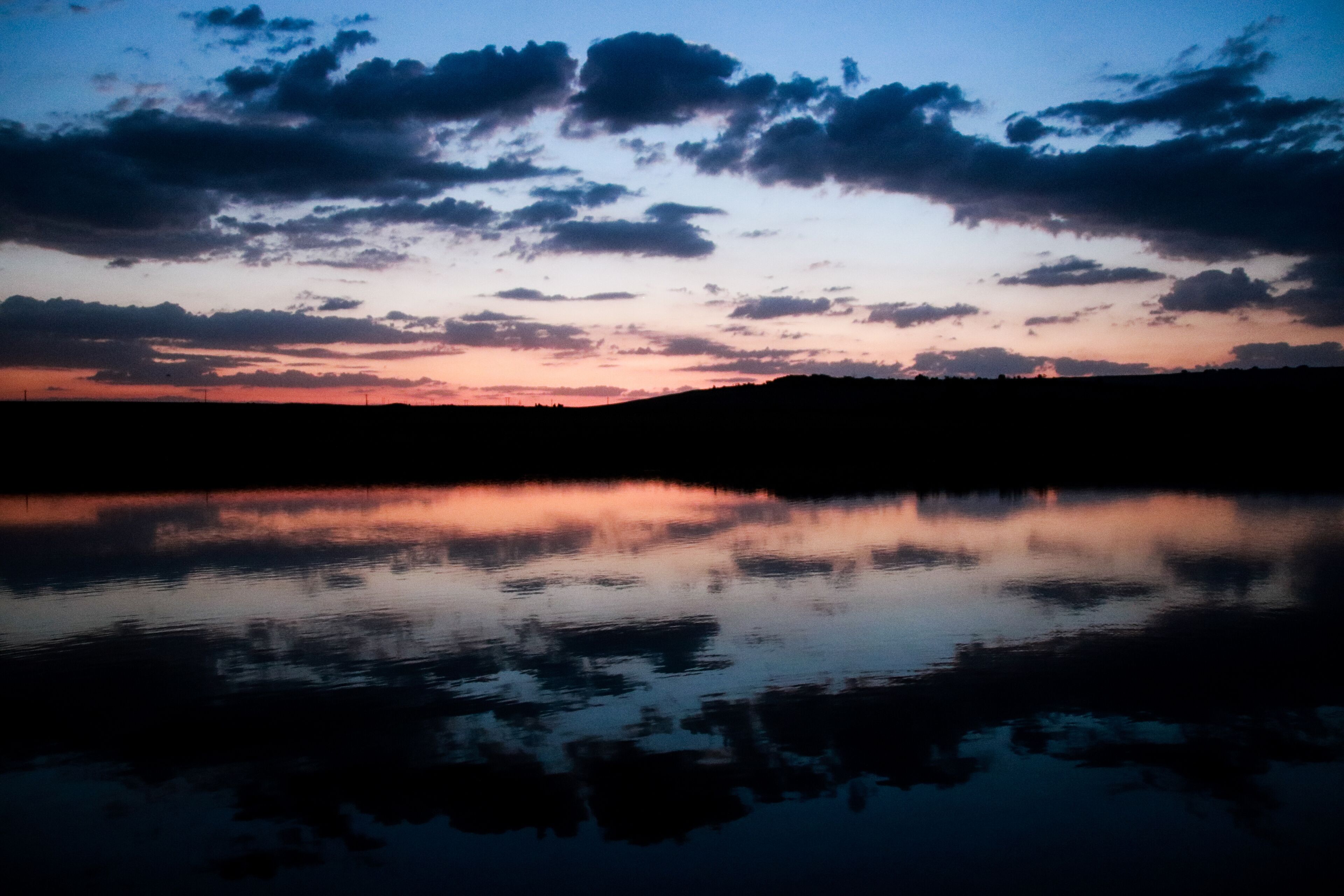 A sunset over a calm lake with silhouetted hills and scattered clouds.