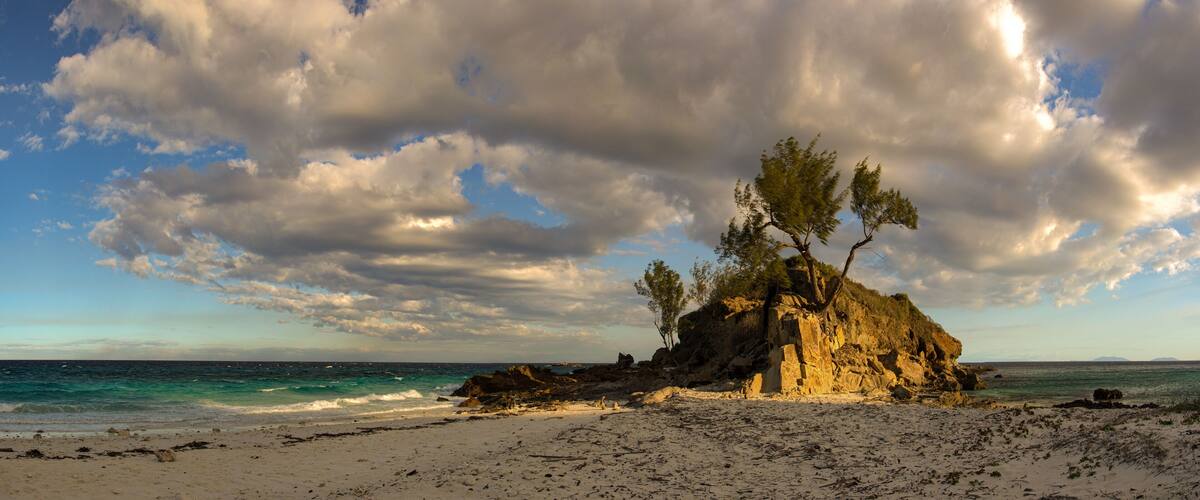 A rocky outcrop with trees on a beach with clear blue water and a cloudy sky.