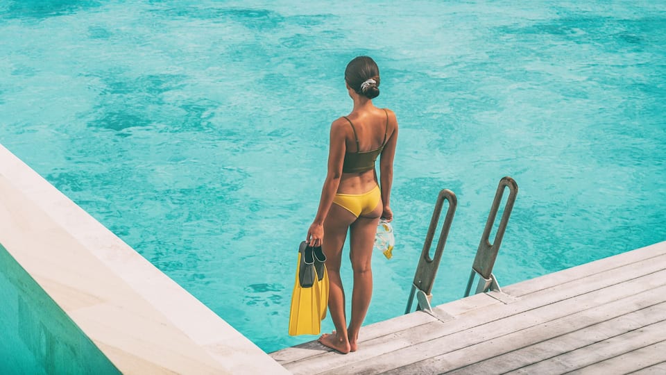 A woman in a yellow swimsuit standing at the edge of a swimming pool with diving boards.