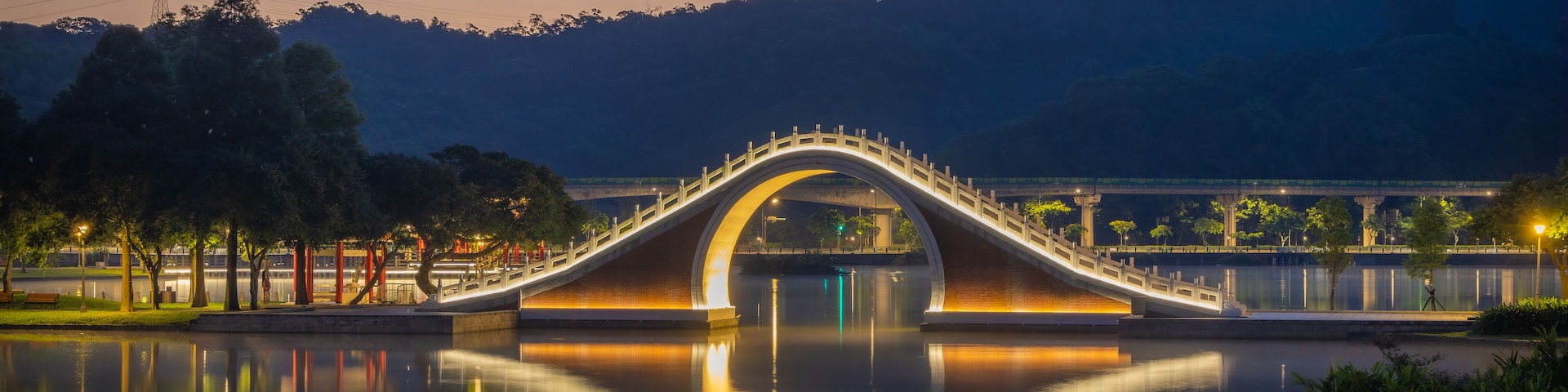 A lit bridge over a calm river at dusk with mountains in the background.