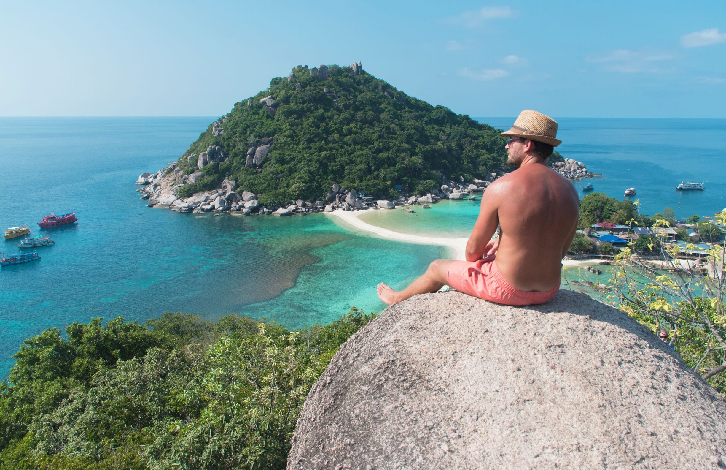 A man sitting on a rock overlooking a turquoise bay with a small island in the background.