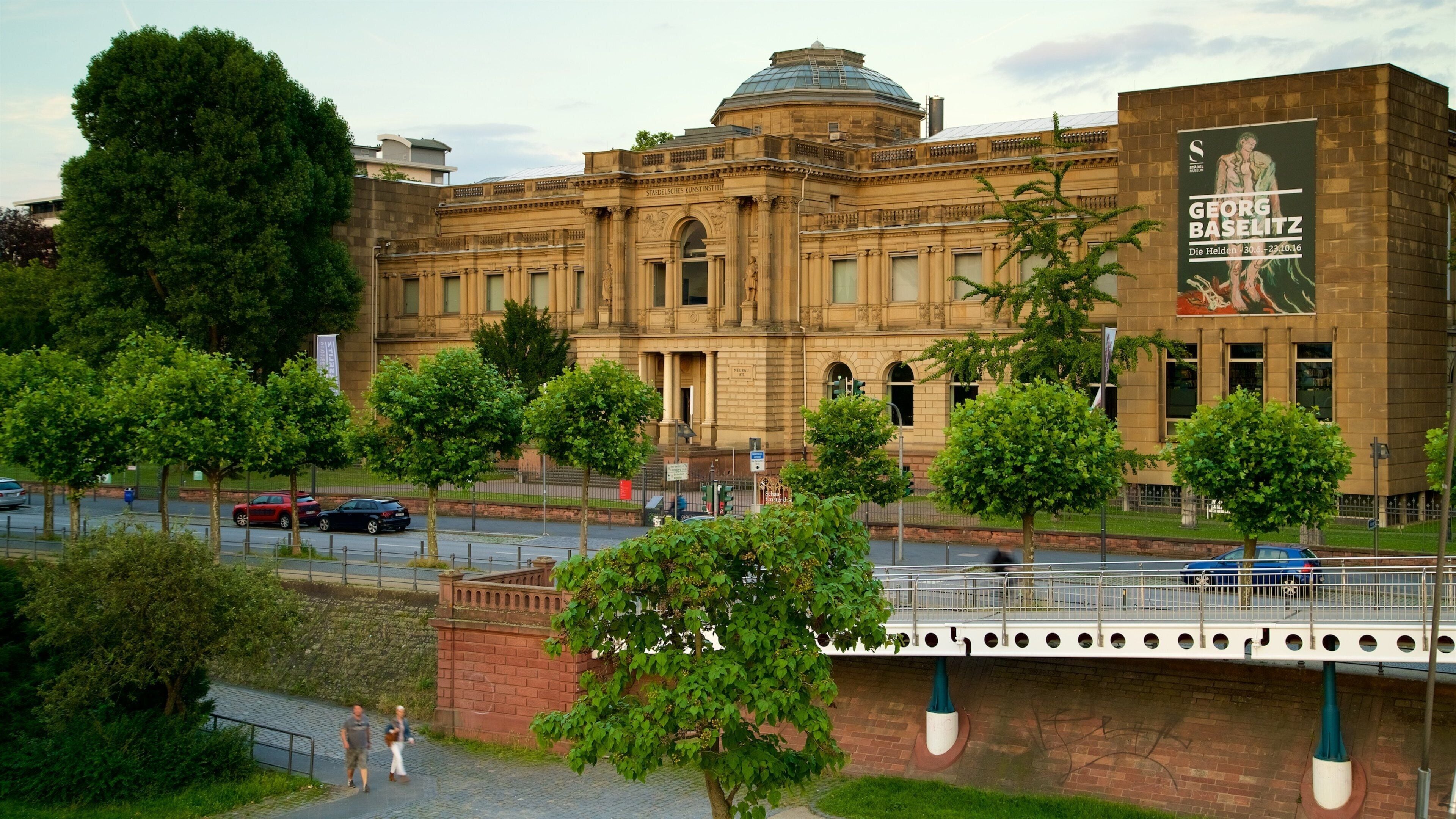 Un edificio histórico con una cúpula y columnas, rodeado de árboles y un puente peatonal.