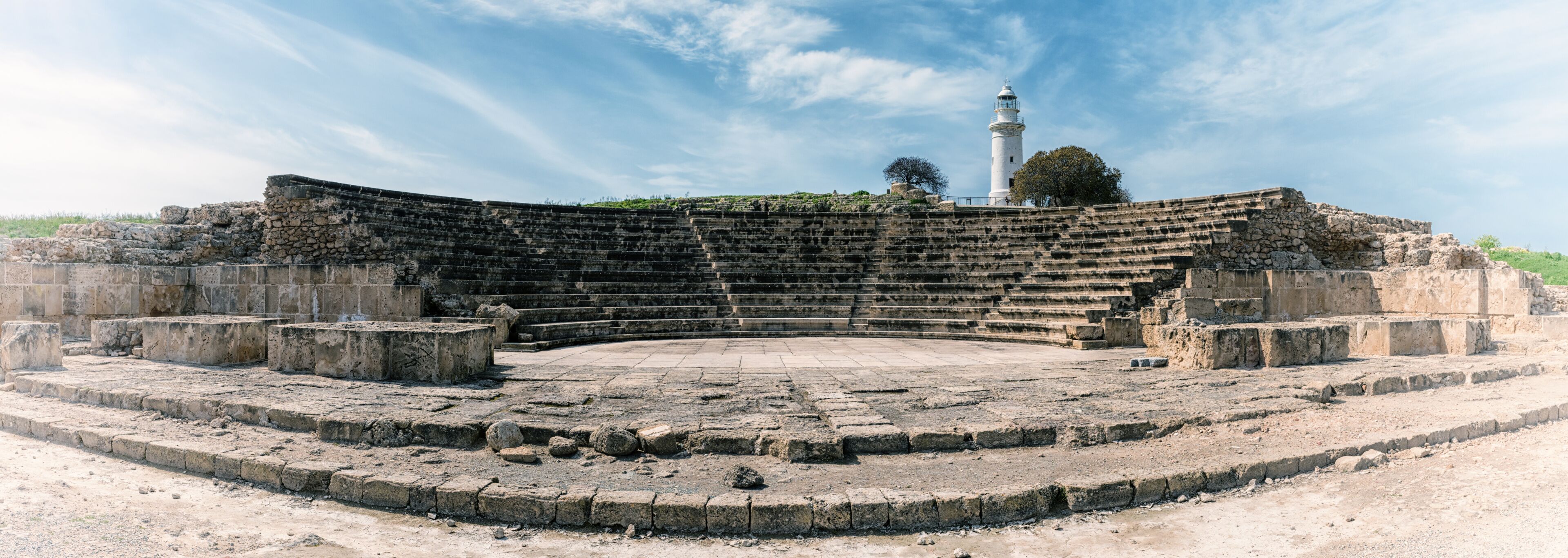 Un anfiteatro antiguo con escalones de piedra y un escenario central, rodeado de ruinas y un faro al fondo.
