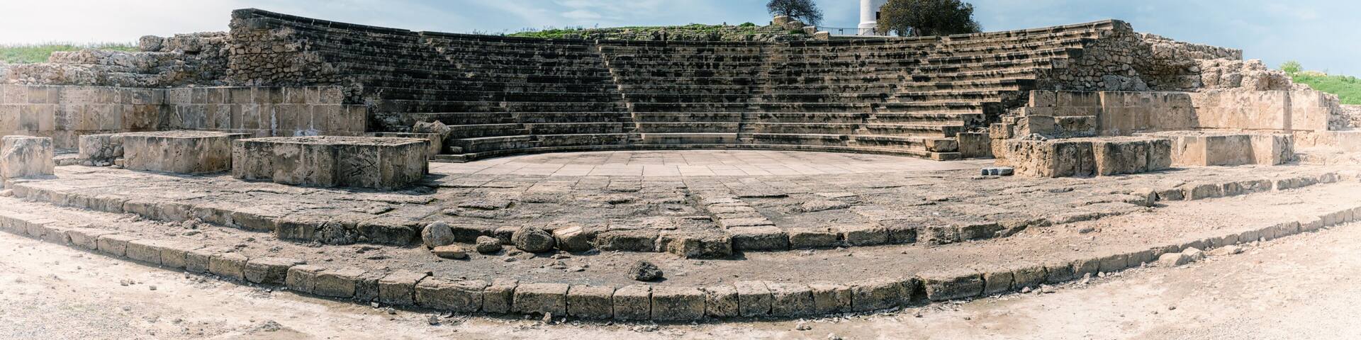 An ancient amphitheater with stone steps and a central stage, surrounded by ruins and a lighthouse in the background.