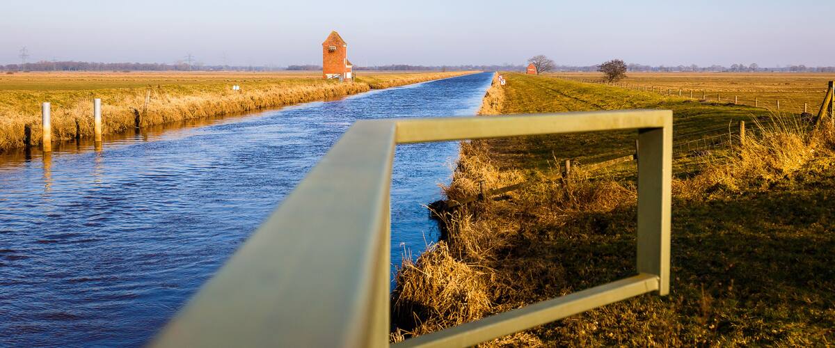 A canal with a wooden frame in the foreground, a red barn in the distance, and open fields.