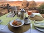 A table set with a meal, including a bowl of snacks, a cup of coffee, and a glass of juice, with a desert landscape in the background.