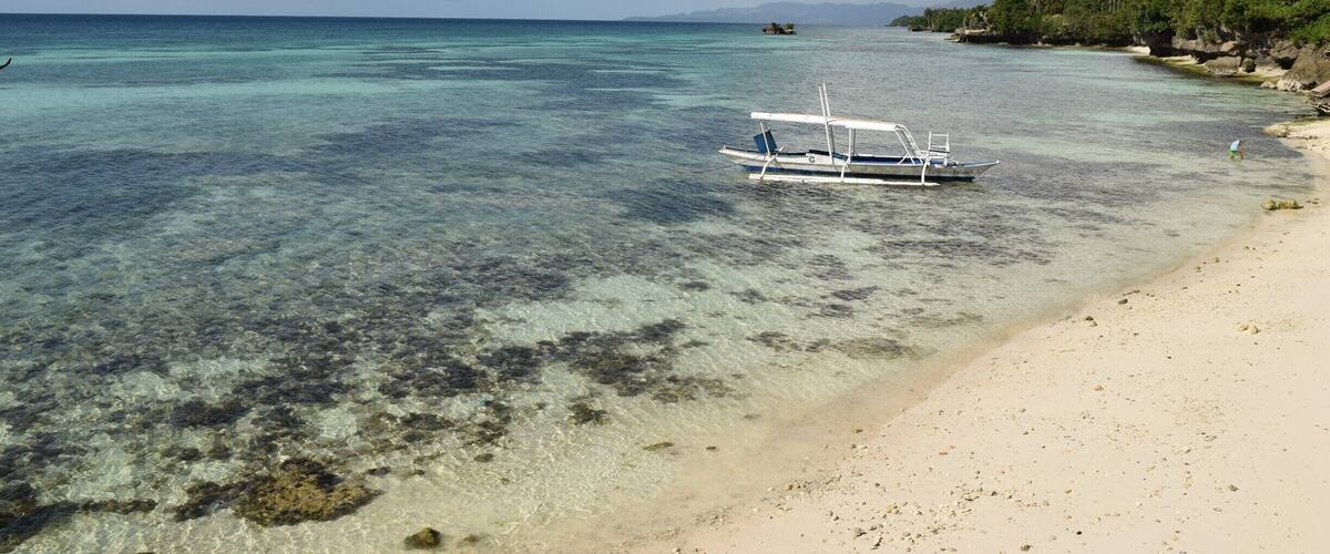 A boat on a clear turquoise sea, a sandy beach, and a forested island in the distance.