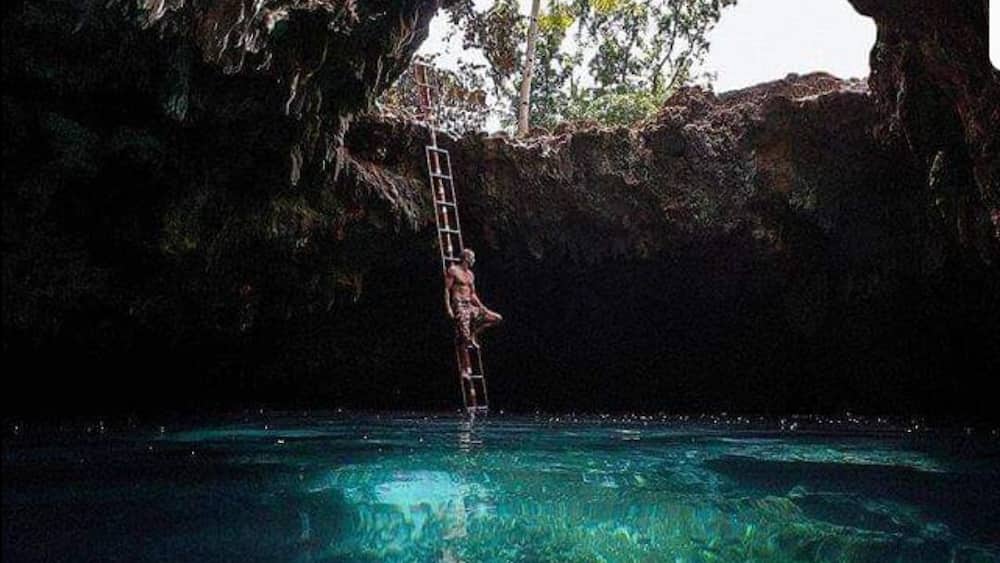 Une personne qui grimpe à une échelle pour se rendre dans une piscine à l’eau cristalline, entourée de falaises rocheuses et de palmiers.