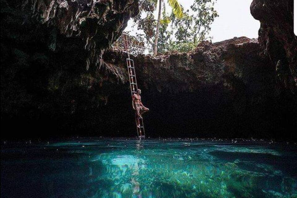 Una persona trepa por una escalera hacia una piscina azul cristalina rodeada de acantilados rocosos y palmeras.