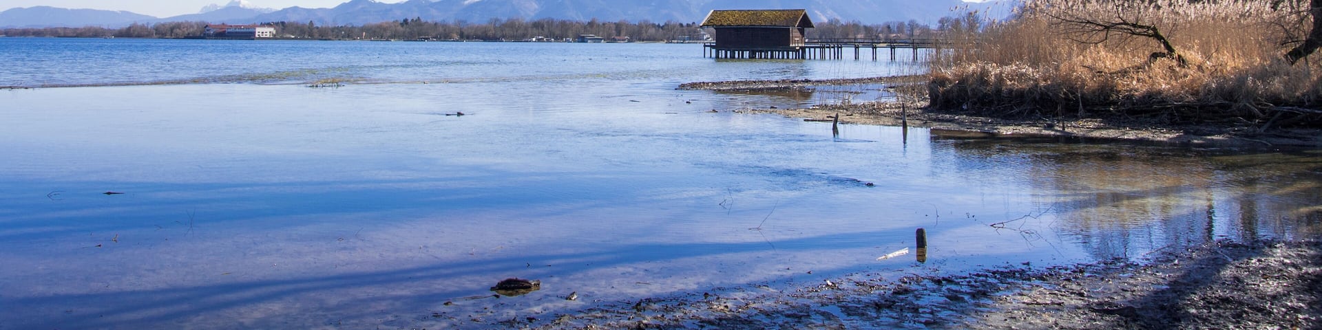 A calm lake with a wooden pier, a house on the shore, and mountains in the background.