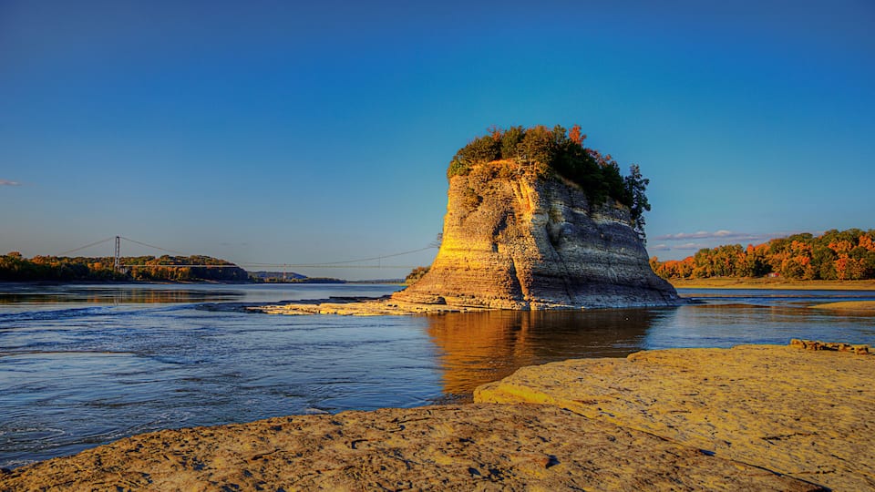 A rock formation by a river with a bridge in the background.