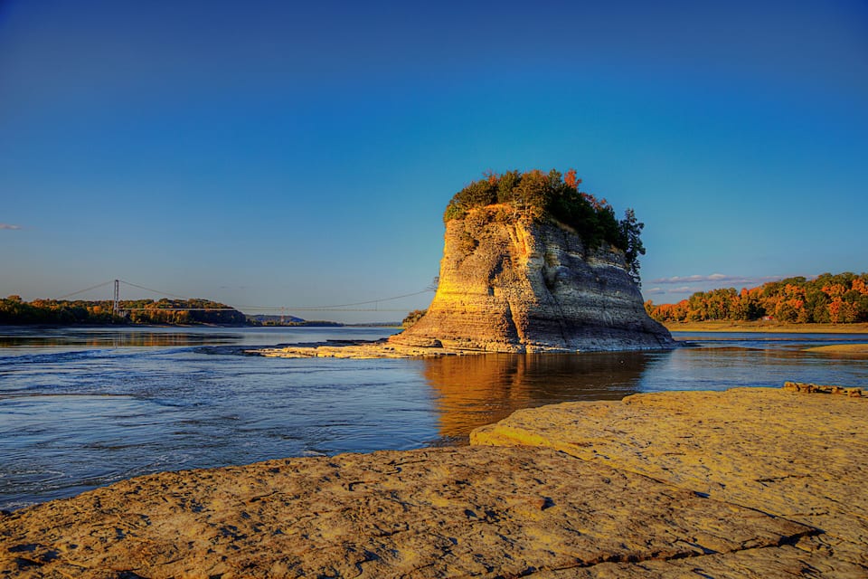 A rock formation by a river with a bridge in the background.
