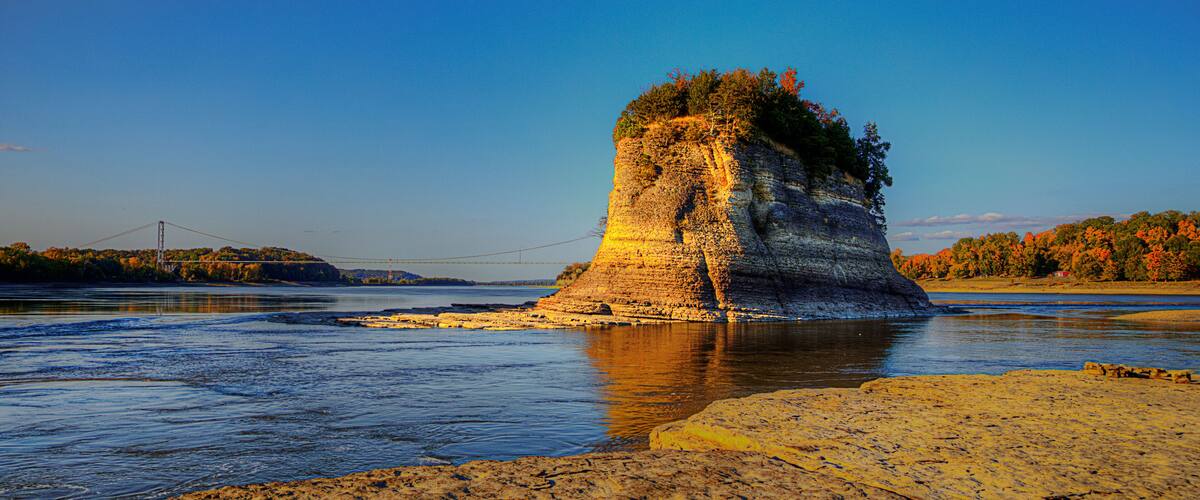 A rock formation by a river with a bridge in the background.