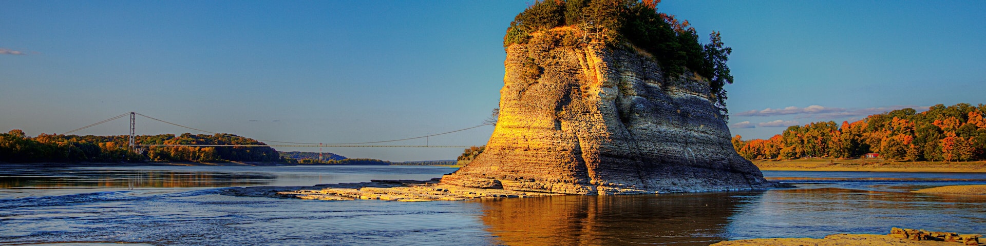 A rock formation by a river with a bridge in the background.