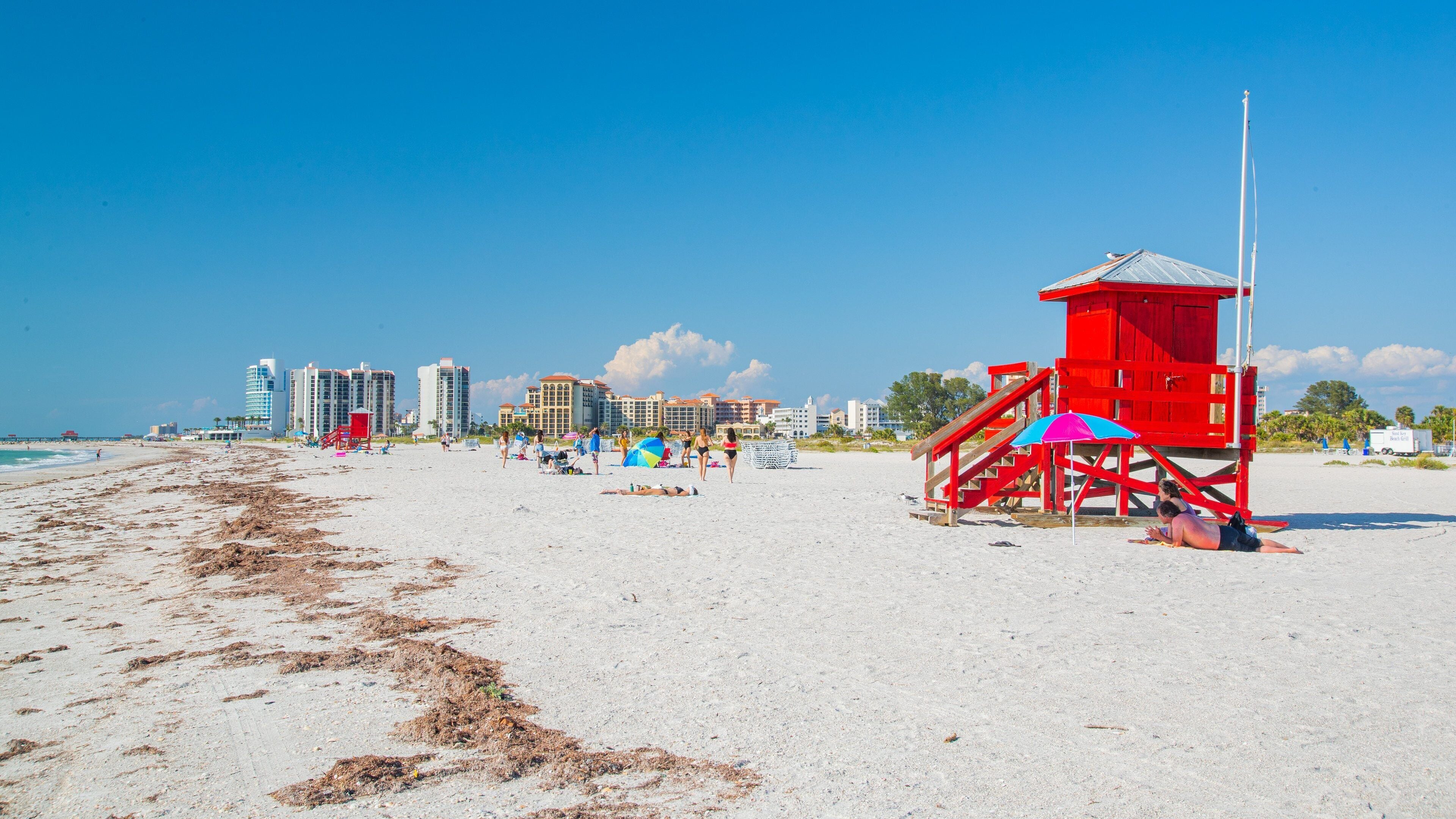 Uma praia com uma torre salva-vidas, guarda-sóis e edifícios ao fundo.