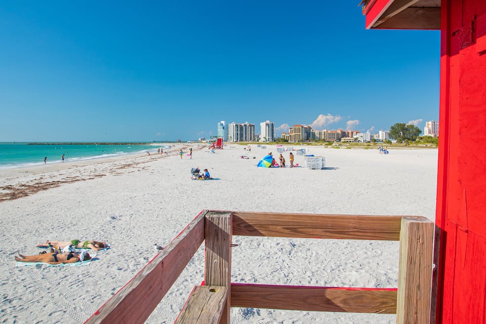 A beach with white sand, a red lifeguard tower, and buildings in the background.