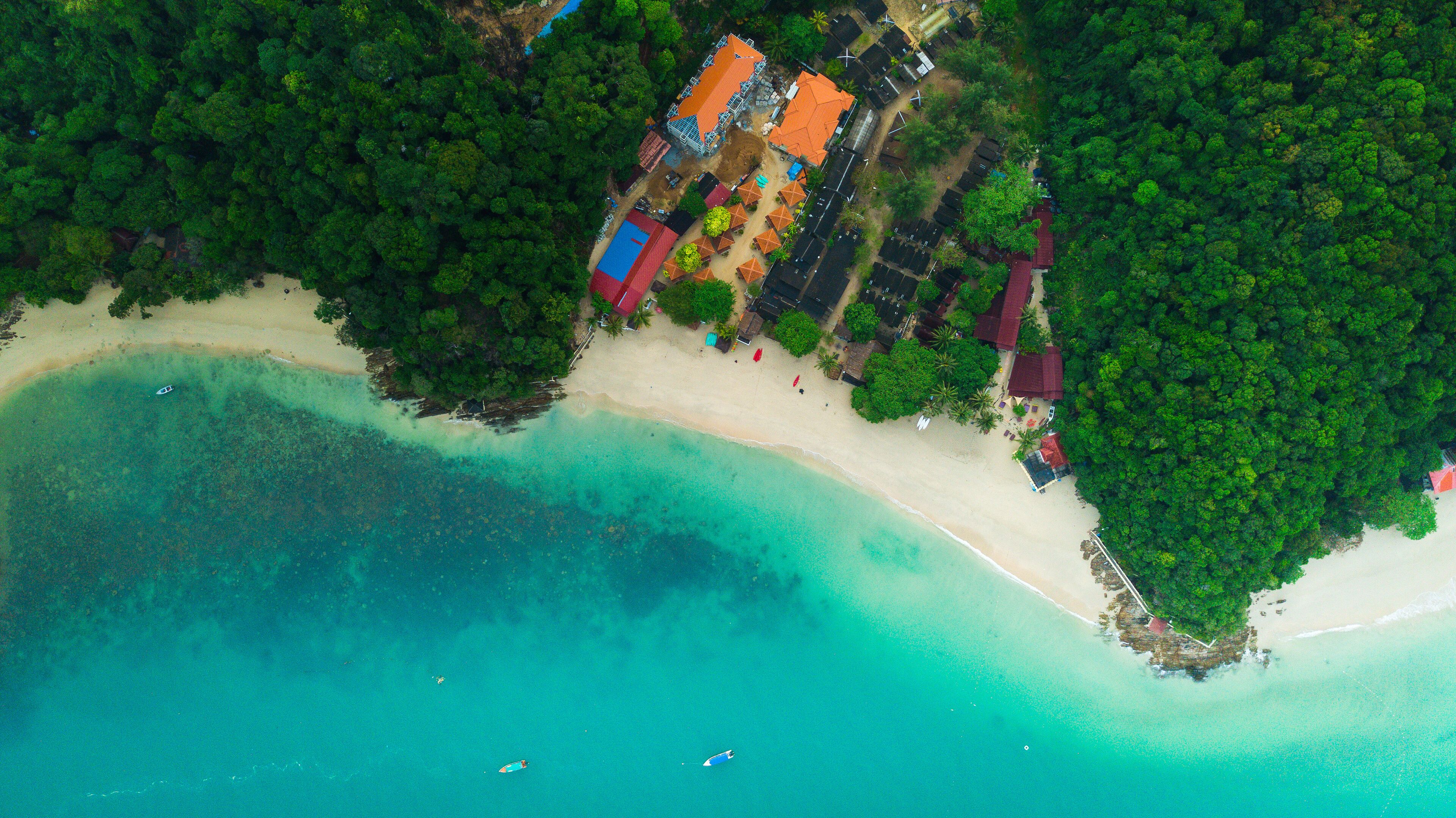 An aerial view of a tropical beach with clear turquoise waters, a sandy shore, and a cluster of colorful buildings surrounded by dense green foliage.