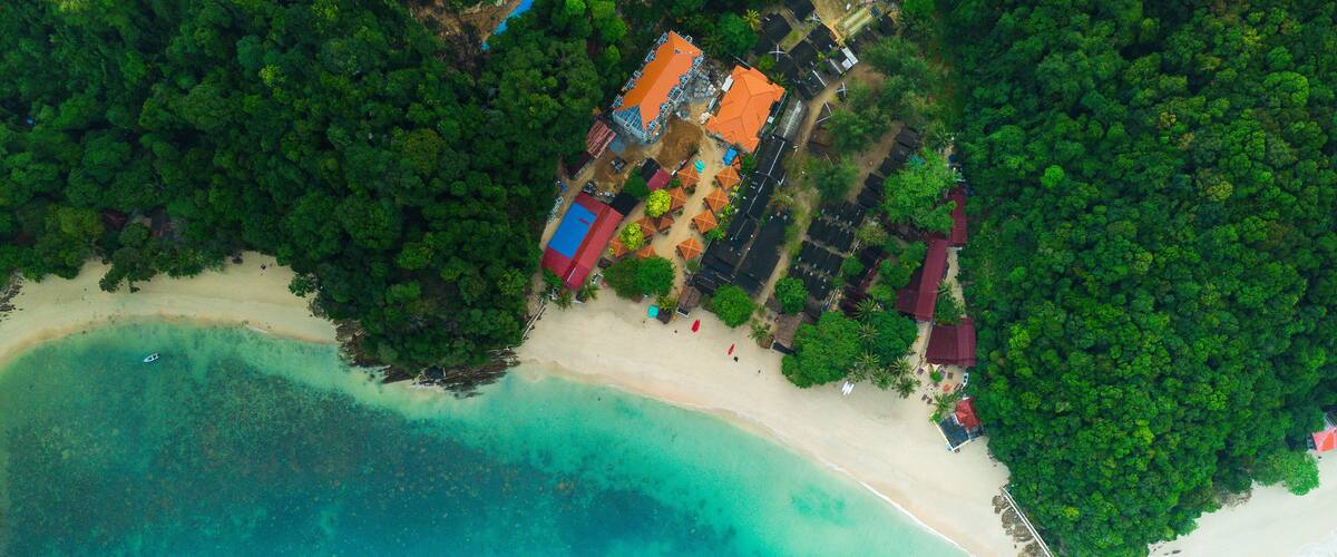 An aerial view of a tropical beach with clear turquoise waters, a sandy shore, and a cluster of colorful buildings surrounded by dense green foliage.
