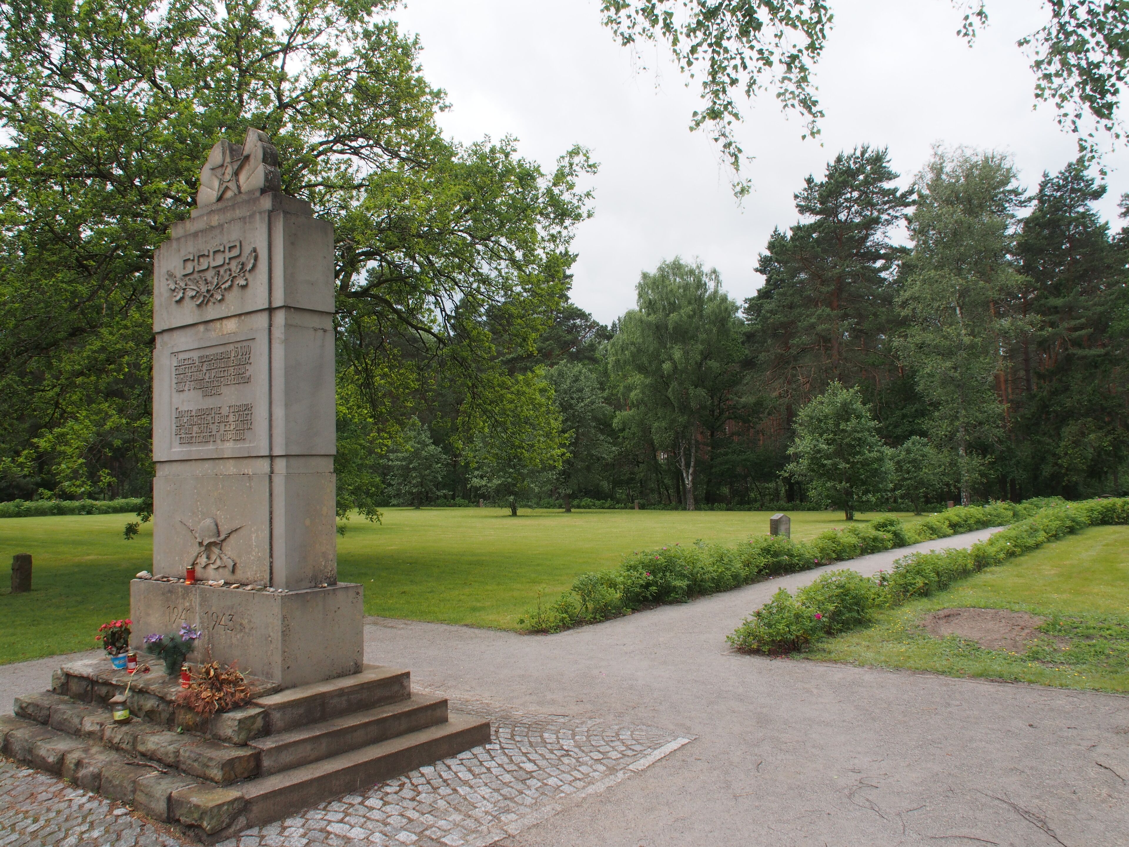 A monument with inscriptions and flowers, surrounded by a park with trees and a pathway.