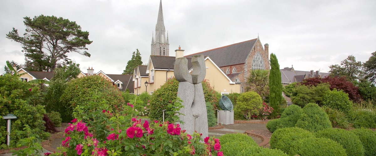A stone sculpture in a garden with vibrant pink flowers, a church with a tall spire, and residential houses in the background.
