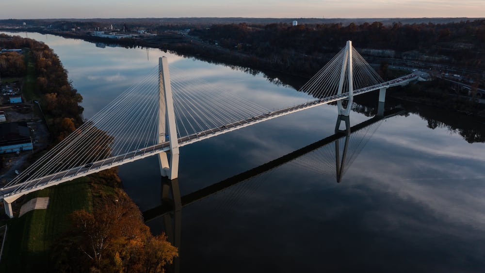 Eine Hängebrücke über einem ruhigen Fluss, umgeben von Herbstlaub und weiter entfernten Gebäuden.
