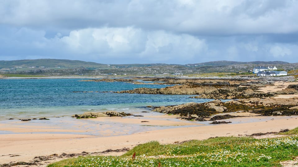 A coastal landscape with a sandy beach, rocky shoreline, and white houses in the background.