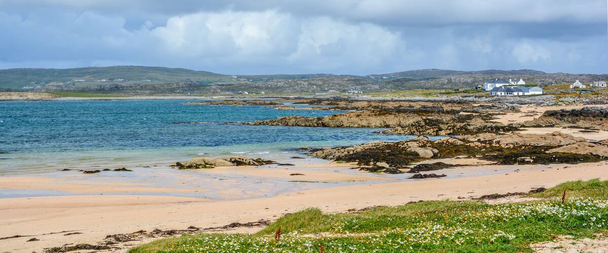 A coastal landscape with a sandy beach, rocky shoreline, and white houses in the background.