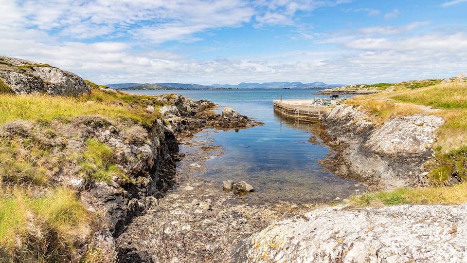 A rocky shoreline with clear water, a pier, and distant mountains under a partly cloudy sky.