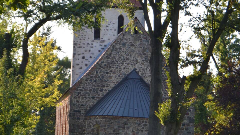 A stone church with a pointed roof and a bell tower, surrounded by trees and grass.