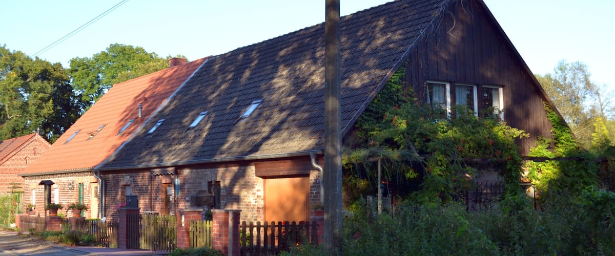 A wooden house with a red roof, surrounded by greenery and a wooden fence.