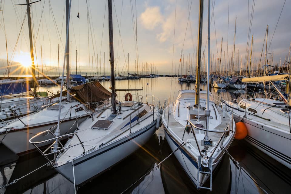 Ein Yachthafen mit mehreren Segelbooten, ein klarer Himmel und eine untergehende Sonne im Hintergrund.