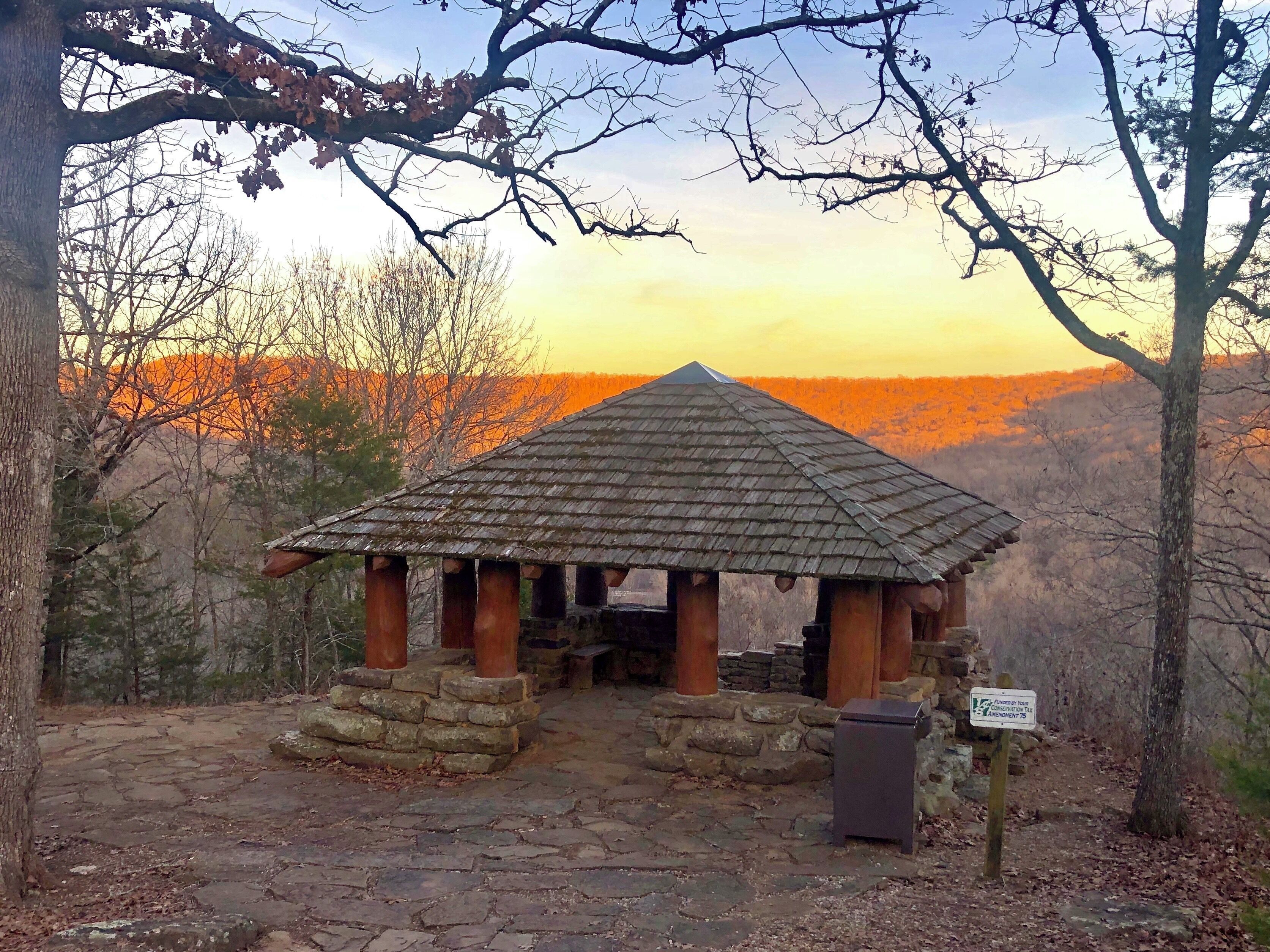A stone gazebo with a tiled roof surrounded by trees and a stone pathway.